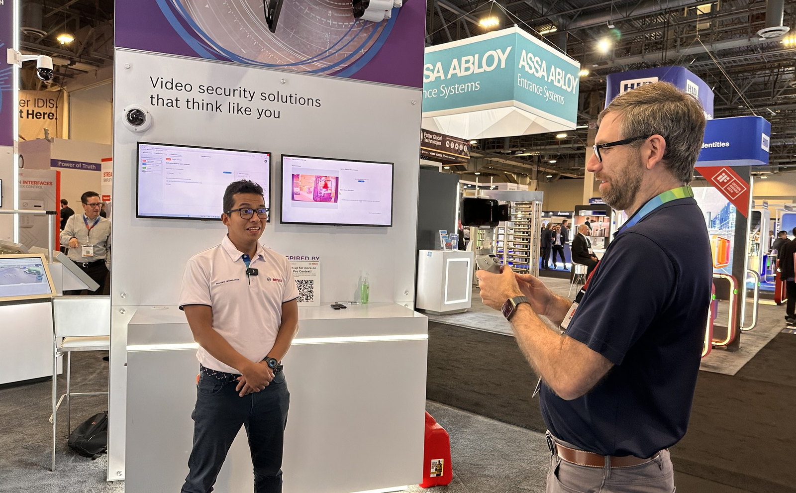 An engineer at a trade show booth being filmed on a phone mounted on a gimbal, a clip mic on his shirt, with the show floor and other stands behind them.